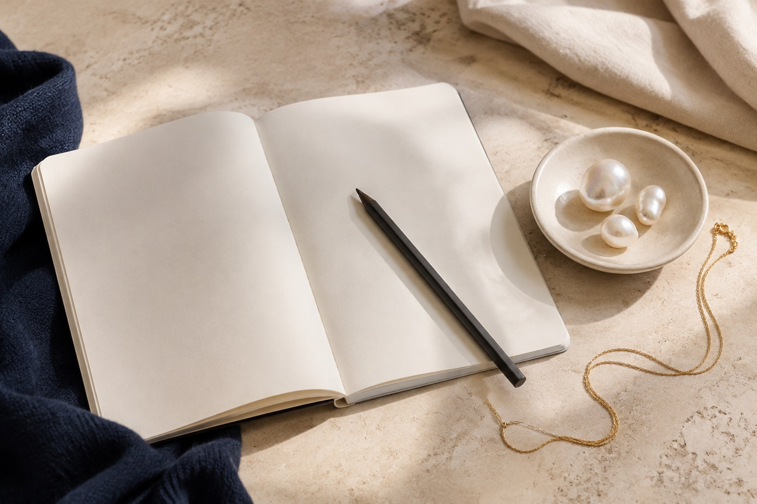 Blank notebook beside three irregular pearls and a fine chain on a warm stone desk.