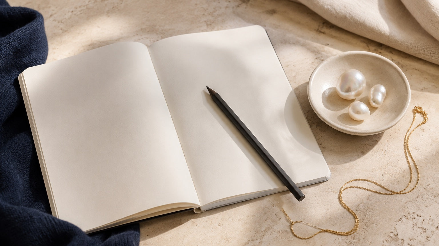 Blank notebook beside three irregular pearls and a fine chain on a warm stone desk.