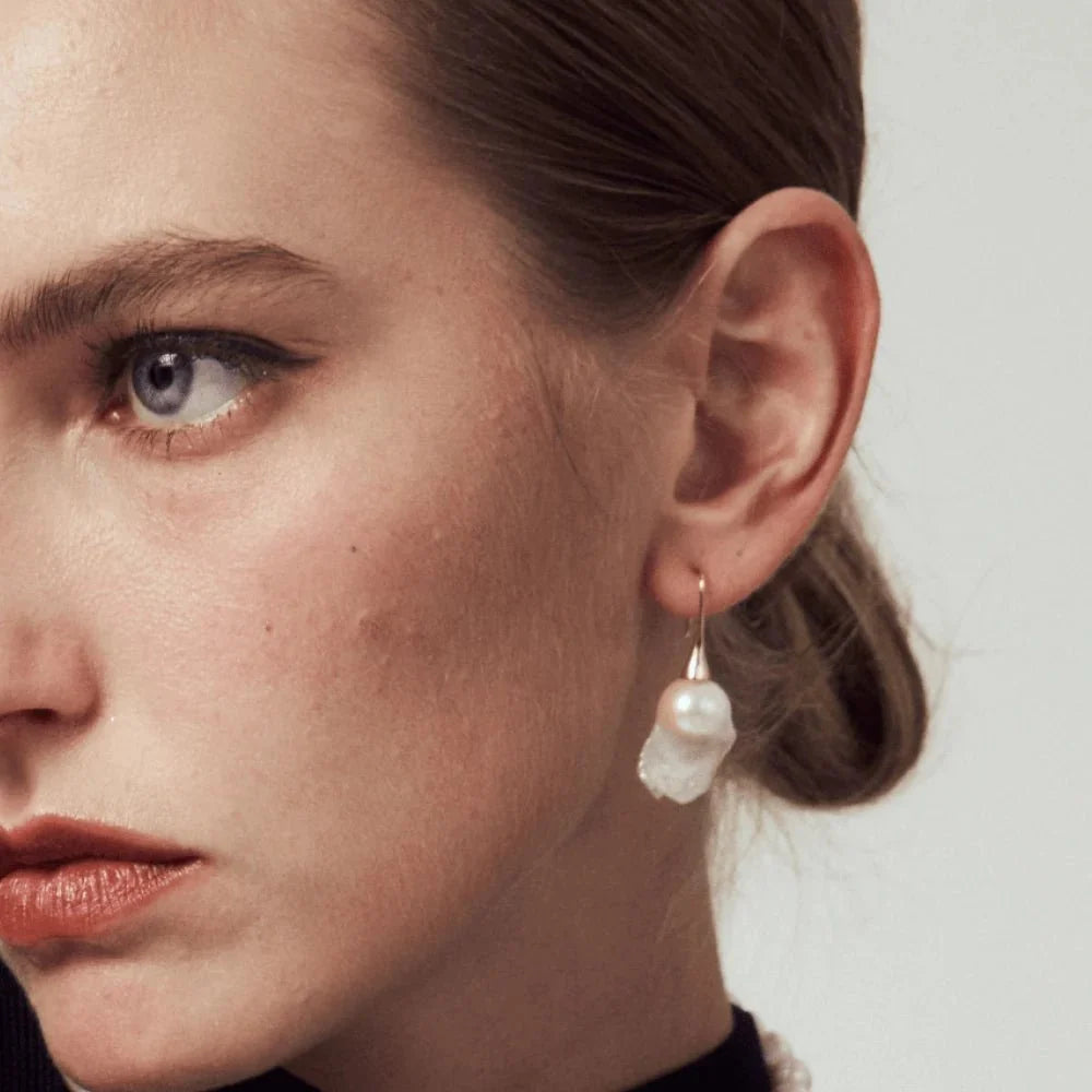 Soft analog film portrait of woman with sleek low bun wearing gold hook baroque pearl drop earrings, natural asymmetrical white pearl with soft iridescent luster