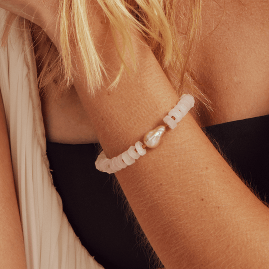 Partial close-up shot of female model’s wrist wearing pink baroque pearl rose quartz bracelet against minimalist black sleeveless soft neutral outfit