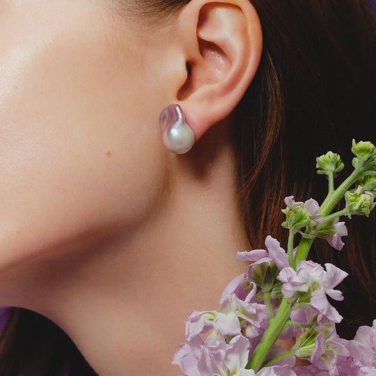 Close-up side shot of female model’s ear, wearing single large purple baroque pearl silver stud earring beside soft purple flower sprigs against pale neutral backdrop