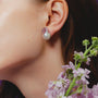 Close-up side shot of female model’s ear, wearing single large purple baroque pearl silver stud earring beside soft purple flower sprigs against pale neutral backdrop