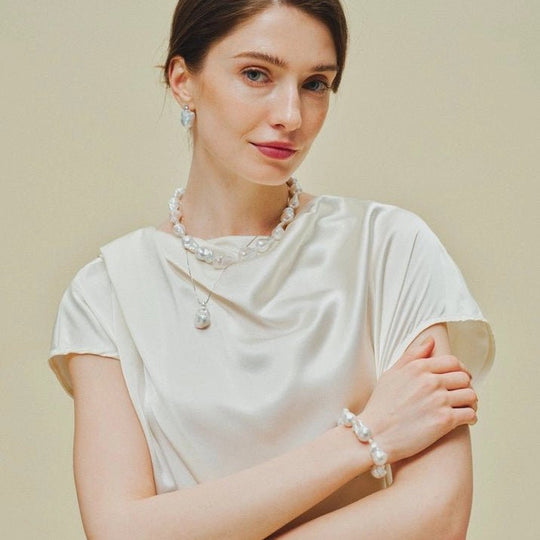 Female model in silky white short sleeve top wearing matching large white baroque pearl earrings, layered necklace and pearl bracelet against soft neutral beige backdrop