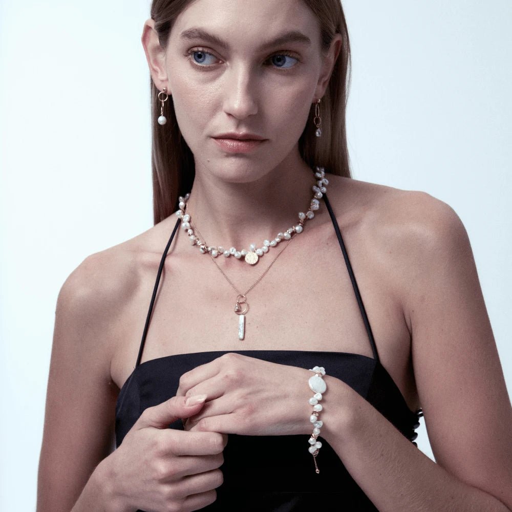 Medium bust shot of woman in minimalist black camisole, layering coin baroque pearl gold bracelet with matching pearl necklaces against soft warm neutral backdrop
