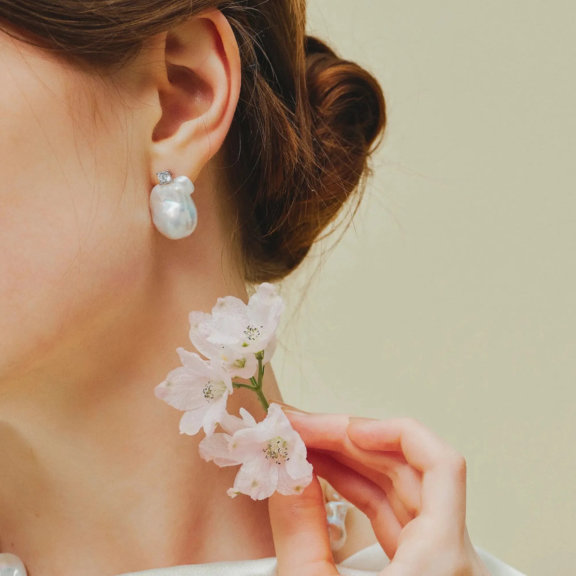 Close-up side shot of woman’s bun hairstyle, single large baroque pearl drop earring paired with small white flower near jaw on muted beige background