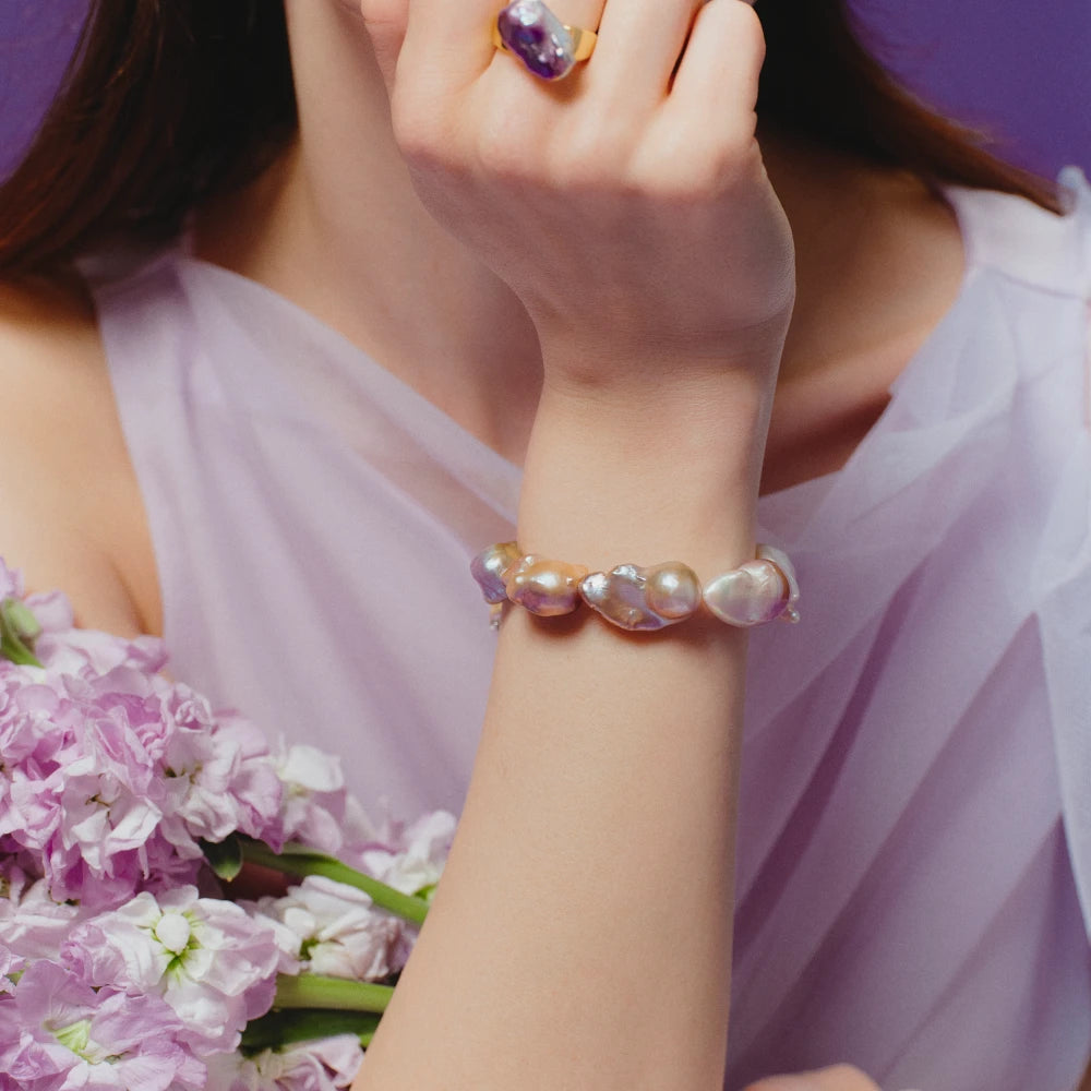 Partial shot of female model’s wrist laid over soft purple flower bouquet, wearing large purple baroque pearl silver beaded bracelet against pale neutral backdrop