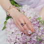 Partial shot of female model’s hand laid over soft purple flower bouquet, wearing adjustable gold purple baroque pearl open ring against pale neutral backdrop