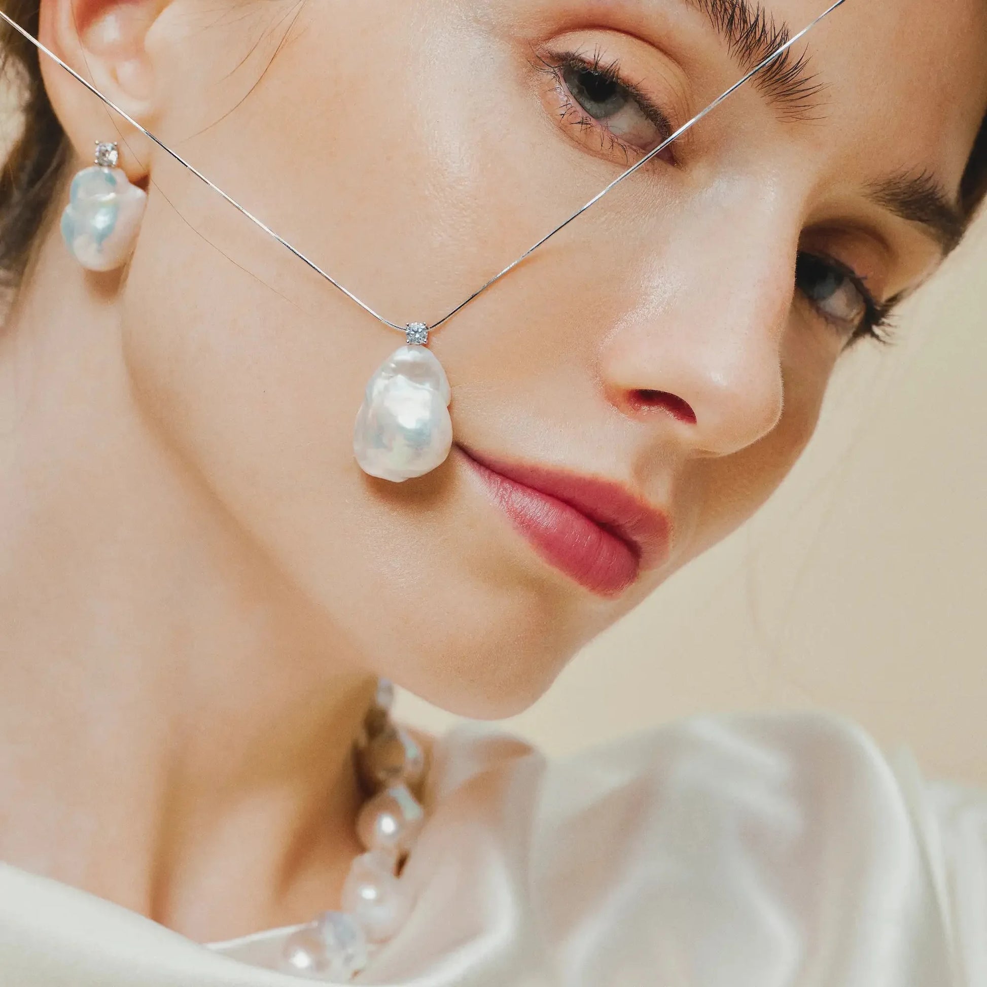 Extreme close-up of female model’s face, showcasing thin silver baroque pearl pendant necklace and matching drop earring against soft plain neutral background