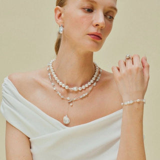 Blonde model in off-shoulder white top, layered with two sterling silver baroque pearl necklaces and matching pearl studs, one hand resting near neck to show stacking effect on soft neutral backdrop