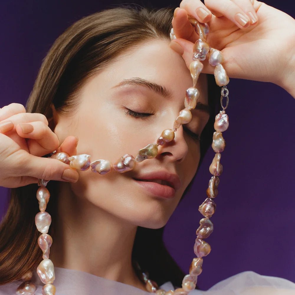 Soft close-up of woman gently holding purple baroque pearl strand necklace, eyes closed against warm dark purple neutral background