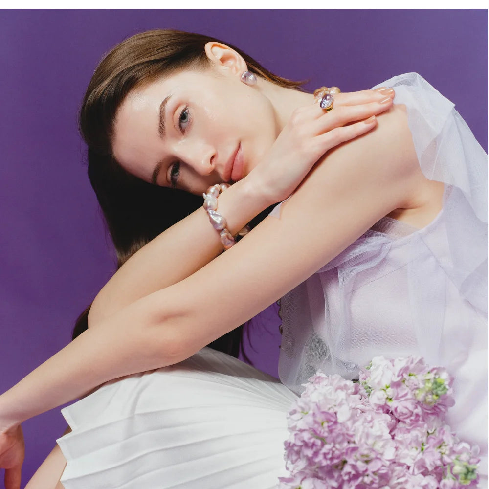 Medium shot of woman in pale sheer camisole, wearing matching purple baroque pearl bracelet and necklace against muted dark purple plain background