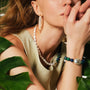 Medium close-up shot of female model’s face and hand, wearing long white oval pearl tassel drop gold earrings beside layered pearl necklaces against soft warm neutral backdrop